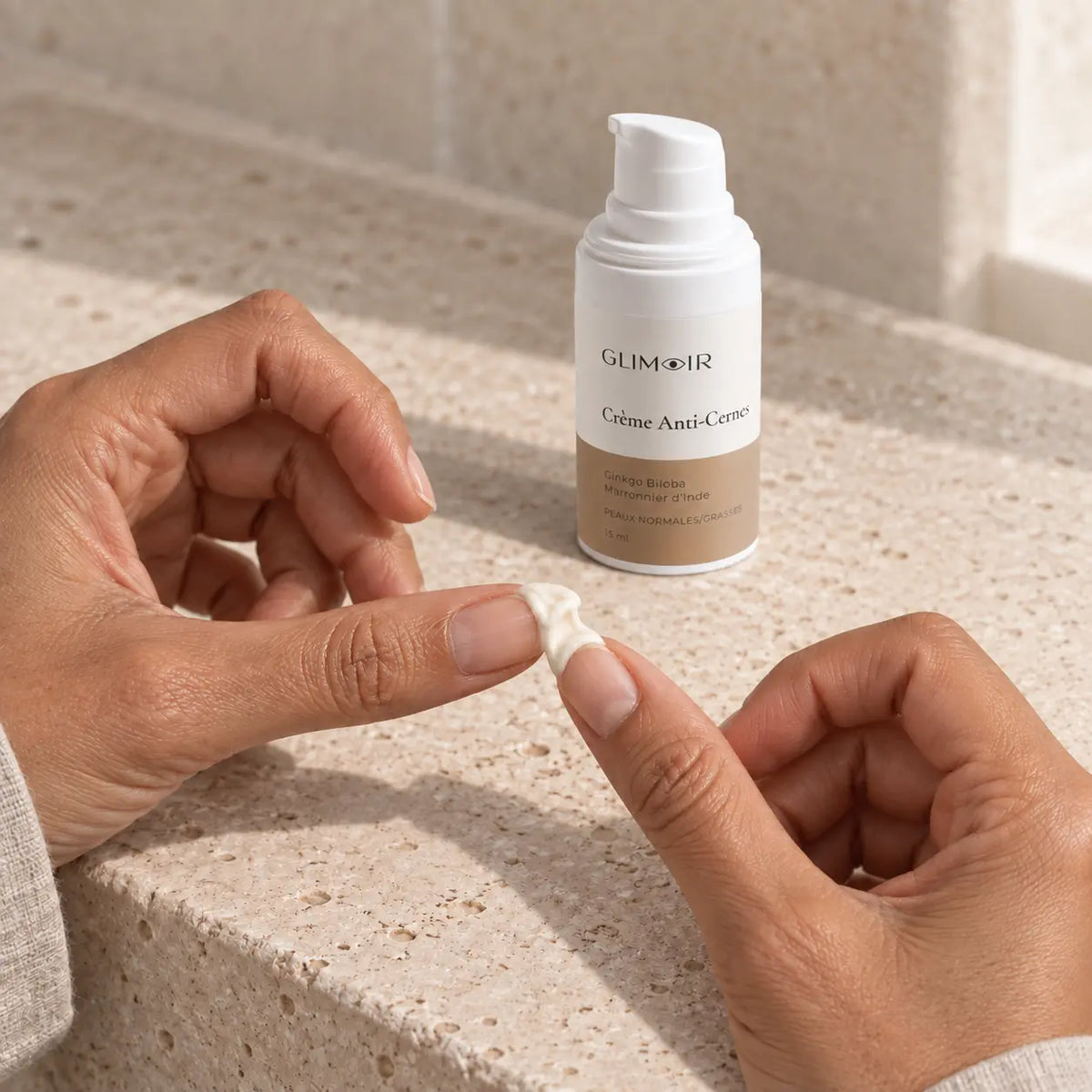 Noisette de Crème Anti-Cernes Glimoir répartie entre les deux annulaires avant application, flacon airless en arrière-plan