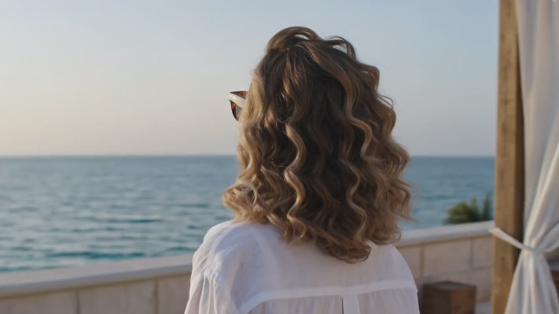 Femme élégante en bord de mer ajustant ses lunettes de soleil, lumière dorée — stick solaire SPF50 regard et visage Glimoir