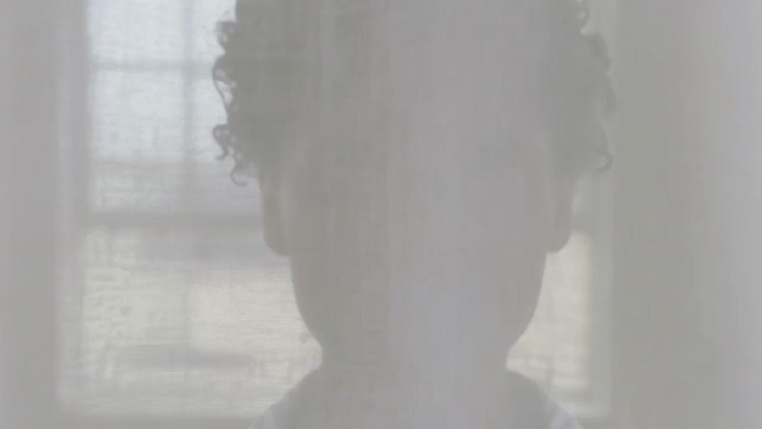 Silhouette d'une femme émergeant d'un voile de rideau vaporeux, yeux fermés, ambiance évanescente associée à la Brume Prébiotique Visage & Regard Glimoir